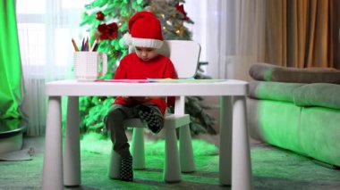 Evde Noel şapkalı bir çocuk var. Şenlik şapkası takan genç bir çocuk arka planda süslenmiş bir Noel ağacıyla masa boyasında oturuyor..