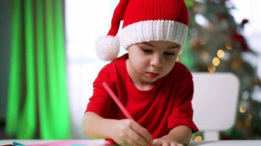 Çocuk şenlik şapkası içinde sanat eseri yapıyor. Kırmızı gömlekli ve Noel şapkalı bir çocuk tatil süslemeleri arasında renkli kalemlerle resim çiziyor..