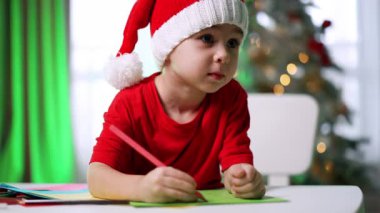 Çocuk rahat bir odada festival sanatı çiziyor. Kırmızı tişörtlü ve Noel Baba şapkalı bir çocuk, dekorasyonlarla çevrili bir masada tatil sanatı çizmeye odaklanmış..