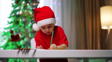 Noel Baba şapkalı çocuk. Noel Baba şapkalı bir çocuk elinde yeşil bir kalemle bir masada oturuyor, süslü bir ağacın yaptığı el sanatlarına odaklanmış..