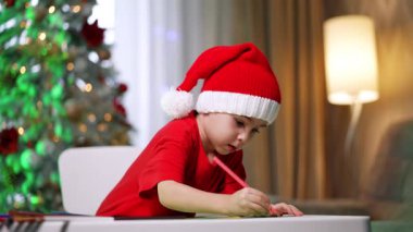 Noel Baba şapkalı çocuk. Noel Baba şapkası takan genç bir çocuk, arka planda süslü bir Noel ağacı bulunan bir masayı boyamaya odaklanır..