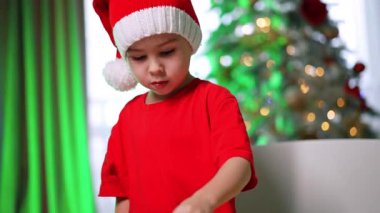 Noel Baba şapkalı çocuk, neşeli bir neşe. Kırmızı tişörtlü ve Noel Baba şapkalı bir çocuk bayram süslerine odaklanıyor..