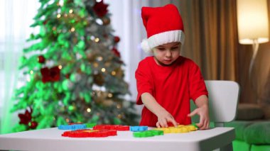Tatil için oyuncakları olan bir çocuk. Kırmızı tişörtlü ve Noel Baba şapkalı bir çocuk süslü bir Noel ağacının yanında renkli bloklarla oynuyor..