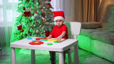 Noel ağacının yanında çocuk oyuncakları. Kırmızı elbiseli bir çocuk bir masada oturuyor, süslü bir Noel ağacının önünde renkli oyuncaklar düzenliyor..