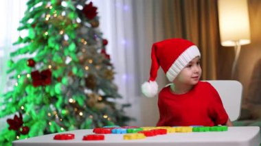 Noel şapkalı çocuk Noel 'in tadını çıkarıyor. Kırmızı tişörtlü ve Noel Baba şapkalı bir çocuk oturma odasındaki süslü Noel ağacının yanında renkli oyuncaklarla oynuyor..