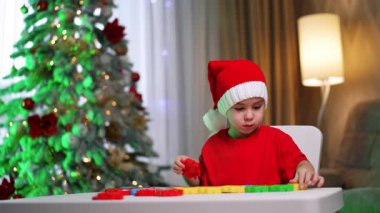 Ağacın yanında çocuk işi. Kırmızı elbiseli bir çocuk renkli şekiller çizerken arka planda süslü bir Noel ağacı parıldıyor..