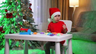 Çocuk içeride tatil dekorasyonu yapıyor. Kırmızı tişörtlü ve Noel Baba şapkalı bir çocuk tatil sezonunda evde renkli bir ağaç süslemesi yapıyor..