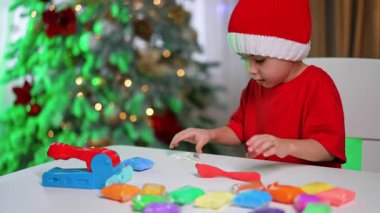 Şenlikte renkli kil eğlencesi. Çocuk, bayram süsleri ve şenlikli bir ağaçla çevrili bir masada renkli killeri neşeyle şekillendirir..