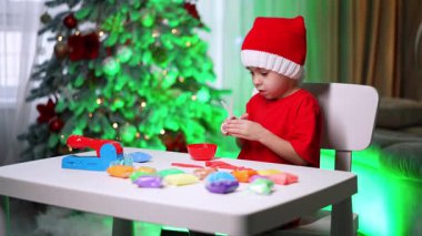 Ağacın orada el işi yapan çocuk. Kırmızı elbiseli ve şapkalı bir çocuk Noel zamanı süslü bir Noel ağacının yanındaki masada el işi yapıyor..