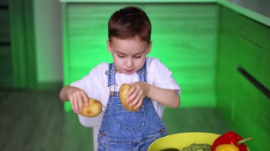 Çocuk içeride sebzeleri keşfediyor. Genç bir çocuk parlak bir mutfakta masada oturmuş, ilginç bir şekilde taze patates ve havuçları inceliyor..