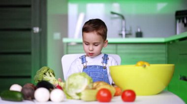 Meraklı çocuk taze sebzeleri inceler. Genç bir çocuk çeşitli taze sebzelerle dolu bir masada oturuyor. Parlak bir mutfakta brokoliyi düşünceli bir şekilde tutuyor..