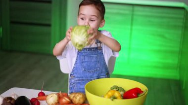 Çocuk mutfaktaki sebzeleri seviyor. Genç bir çocuk elinde lahanayla gülümsüyor, parlak bir mutfakta renkli sebzelerle çevrili..