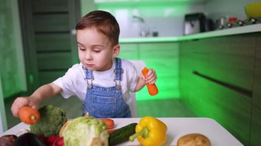 Çocuk içeride sebze keşfediyor. Kot pantolon giymiş bir çocuk havuç tutarken, çeşitli sebzelerle çevrili, parlak bir mutfakta..