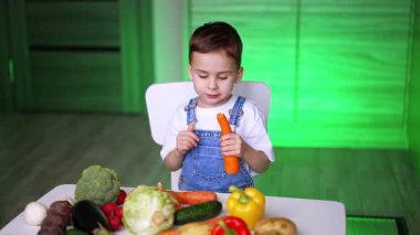 Parlak mutfakta sebze doğrayan çocuk. Genç bir çocuk, çeşitli taze sebzelerle dolu bir masada oturmuş renkli bir mutfakta havuç tutmaya odaklanmış..