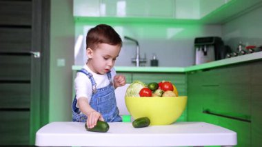 Çocuk mutfaktaki sebzeleri keşfediyor. Tulumlu bir çocuk sağlıklı beslenmeyi öğrenirken parlak bir mutfakta çeşitli taze sebzelere dokunur..