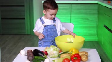 Çocuk yemek pişirmek için sebze hazırlar. Mavi tulumlu küçük bir çocuk mutfak masasındaki büyük sarı bir kaseye taze sebze koyar..