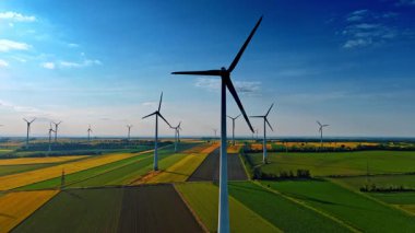 Türbinler yenilenebilir enerji üretir. Rüzgâr türbinleri renkli tarlaların üzerinde, kırsal alandaki parlak mavi gökyüzünün altında rüzgar enerjisini dizginler..