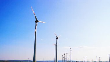 Gündüz temiz enerji türbinleri. Rüzgâr türbinlerinden oluşan bir hat açık mavi bir gökyüzüne karşı duruyor. Gün boyunca güneş ışığı kılıçlarını aydınlatıyor..