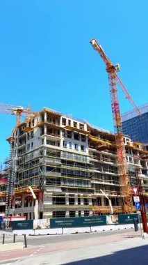 Kentsel alanda yeni bir inşaat projesi. İşçiler kalabalık bir şehirde, açık mavi gökyüzünün altında vinçleri ve iskelesi olan büyük bir bina inşa etmekle meşguller..