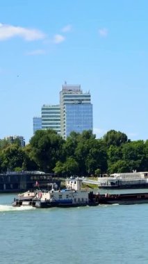 Şehrin ufuk çizgisine yakın tekneleri olan bir nehir kenarı. Tekneler nehir kıyısında demirli ve güneşli bir günde arka planda modern bir şehir silueti var..