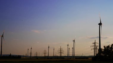 Güneş batarken gökyüzü çok güzel. Yel değirmenleri ve elektrik kuleleri var. Enerji üretimi ve iletim kavramı.
