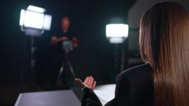 Tanımlanamayan esmer kadın kameraya arkasını döner ve eliyle konuşur. Kameraman sahne arkasında çalışırken bulanık arkaplanda görülebilir.