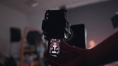 Uzun esmer, uzun saçlı, siyah ceketli ve beyaz gömlekli bir kadın. Erkek el kamerayı tutuyor. Sahne arkasında video kaydı. Kapat..