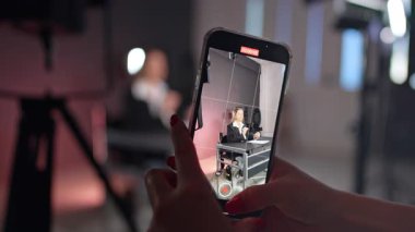 Kırmızı manikürlü kadın eller, masada oturan ve konuşan bir kadının videosunu çekerken telefonu tutun. Modern stüdyoda bir blog görüntüsü.