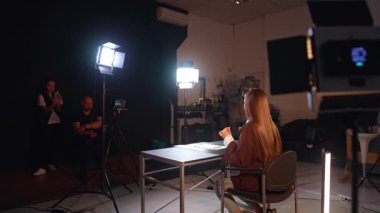 Kahverengi ceketli uzun saçlı esmer kadın modern fotoğraf stüdyosunda kameraya yarım yamalak oturuyor. Erkek fotoğrafçı ve kadın asistan sahne arkasında çalışıyor..