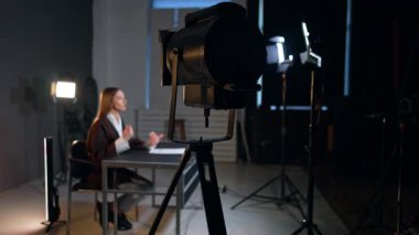 Soffits kameraya konuşan kadın muhabiri aydınlatıyor. Kahverengi ceketli kadın masada oturuyor ve ellerini sallayarak konuşuyor..