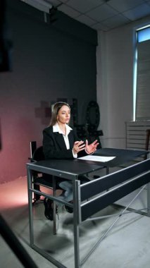 Siyah ceketli esmer kadın stüdyoda masasında oturuyor. Kadın bir blog ya da podcast çekerken kamerayla konuşuyor. Dikey video.