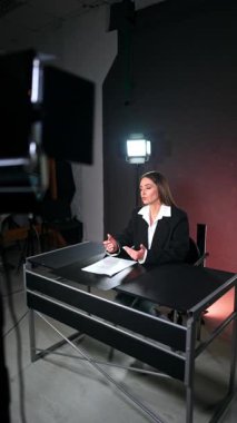 Uzun esmer, siyah ceketli güzel beyaz kadın stüdyoda masasında oturuyor. Soffit 'ler kadın blogcuya yöneliktir. Dikey video.