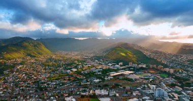 Honolulu, Hawaii 'nin yeşil dağlarla çevrili şehirleri. Güneş ışınları dramatik bulutların içinden geçer. Hava görünümü.