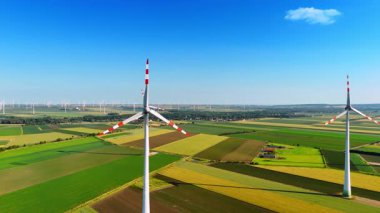 Tarlalarda uzun rüzgar türbinleri. Rüzgâr türbinleri, açık mavi gökyüzünün altında, geniş yeşil bir arazide huzur içinde dönerler..