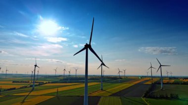 Açık mavi gökyüzü altında geniş bir rüzgar çiftliği. Güneşli bir arazide rüzgâr türbinleri yükselir ve tarım arazilerindeki yenilenebilir enerjiyi vurgular..
