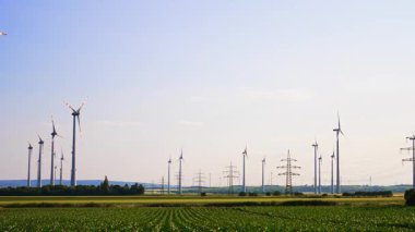 Rüzgar türbinleri ve elektrik destekli tarlalar. Arka planda bir uçakla açık mavi gökyüzü. Düşük açı görünümü.