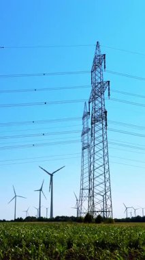 Rüzgar türbinleri ve elektrik hatları. Rüzgâr türbinleri tarlada dönerken enerji hatları güneşli bir günde açık mavi gökyüzüne karşı yükseliyor..