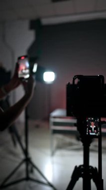 Tripodun üzerindeki kamera bir kadınla video kaydediyor. Yanında duran tanınmayan kadın telefonuyla video çekiyor. Arkaplan bulanık. Dikey video.