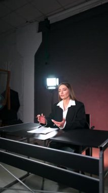 Uzun esmer saçlı, beyaz gömlekli ve siyah ceketli bir kadın masada oturuyor. Blogcu kameraya el sallayarak konuşuyor. Video kaydının kulisinde. Dikey video.