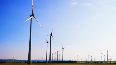 Güneşli bir günde türbinler. Rüzgâr türbinleri, açık mavi bir gökyüzü altında kırsal alanda yavaşça dönerek yenilenebilir enerjiyi iş başında gösteriyor..