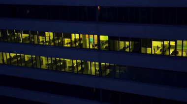 Multi-storied office building with desks and tables. Dark time footage outside the building with light switched on inside.