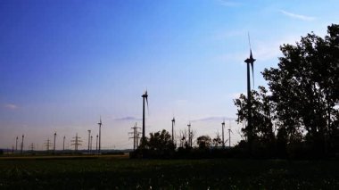 Rüzgar türbinleri günbatımında enerji üretir. Rüzgar türbinleri, açık yeşil bir arazide yenilenebilir enerjiyi kullanan renkli bir günbatımına karşı dimdik dururlar..