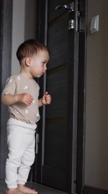 Sevimli çocuk kapının yanında duruyor. Çocuk kapıyı itiyor ve nasıl açıldığına bakıyor. Düşük açı. Dikey video