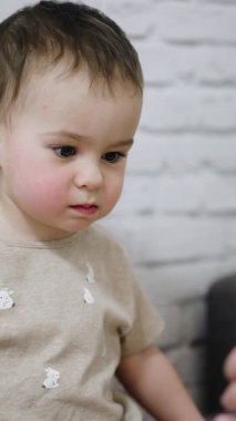 Sevimli küçük çocuk telefonda gösterilen bir şeyle eğleniyor. Sevimli bebek gülümser ve tatlı tatlı kıkırdar. Beyaz duvar arkaplanı. Dikey video