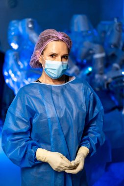 Robotlu cerrahi personel. Maskeli bir sağlık görevlisi mavi ışıklı bir odada duruyor. Yanında da robotik cerrahi asistanları var..