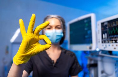 Tıbbi uzman onay işareti veriyor. Koruyucu dişli bir sağlık görevlisi, klinik ortamda sarı eldivenli bir elle onaylama sinyali veriyor..