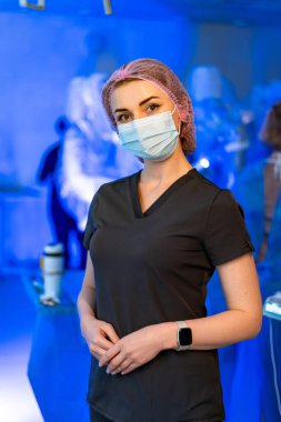 Klinik sağlık uzmanı. Önlük giymiş bir sağlık çalışanı mavi ışıklandırılmış bir tıbbi ortamda maske takarak güvenle ayakta duruyor..