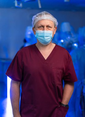 Cerrahi giyimli sağlık çalışanı. Önlüklü ve maskeli bir adam, arka planda profesyonellerin çalıştığı tıbbi bir ortamda güvenle duruyor..