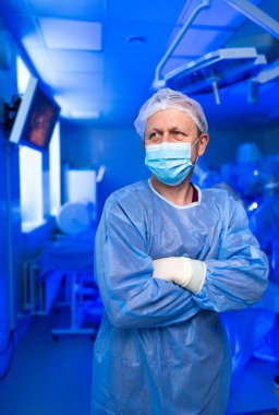 Ameliyathanede tıp profesörü. Tam koruma teçhizatı olan bir tıbbi uzman ameliyat sırasında mavi ışıklı bir ameliyathanede kolları çapraz duruyor..