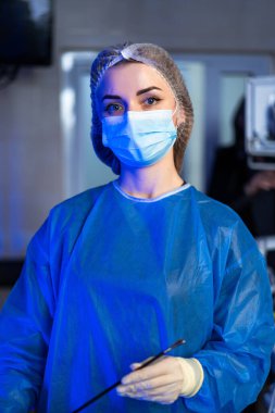 Cerrah ameliyata hazırlanıyor. Ameliyat odasında önlük ve maske takmış bir sağlık çalışanı hazır bekliyor. Hastanın güvenliğini ve bakımını sağlıyor..